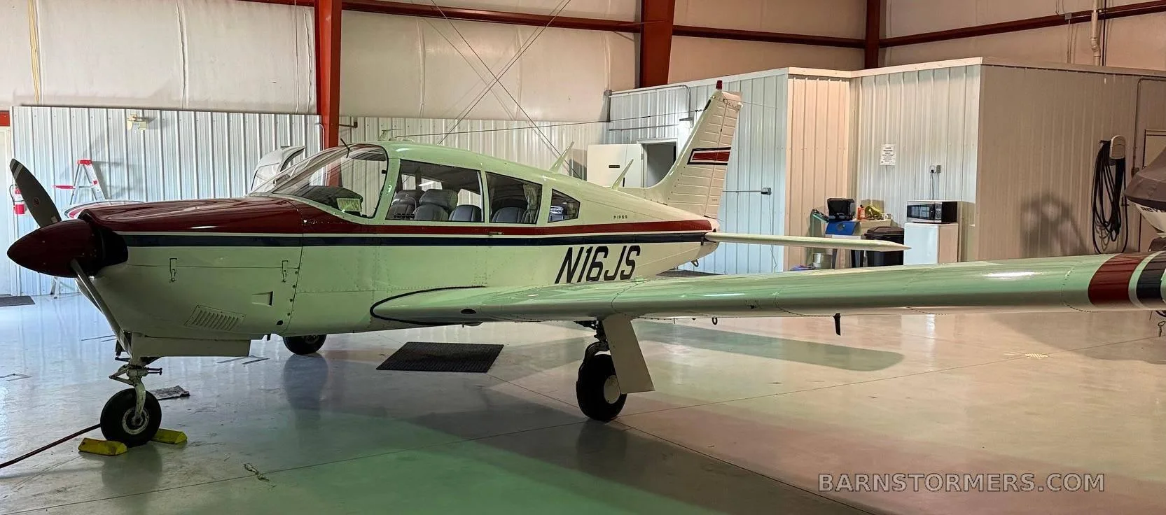 A white Piper aircraft with red and blue stripes, registration N16JS, parked inside a hangar.