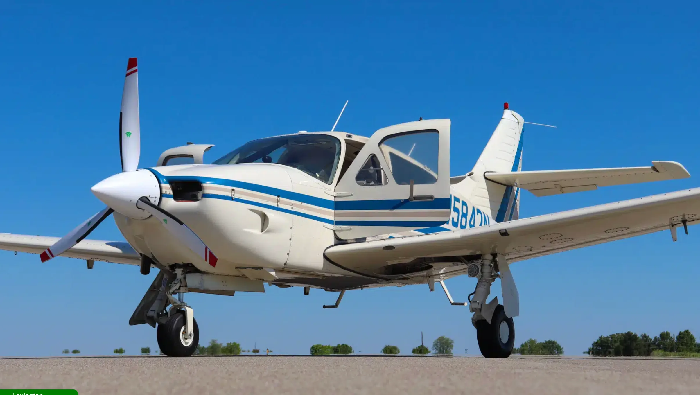 Low-angle front-left view of a white single-engine propeller aircraft with blue and tan stripes, parked on an airfield with doors open.