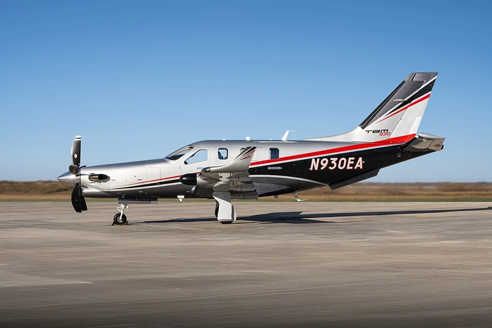 A silver and black Daher TBM 930 turboprop aircraft with red and white accents is parked on a tarmac under a clear blue sky.
