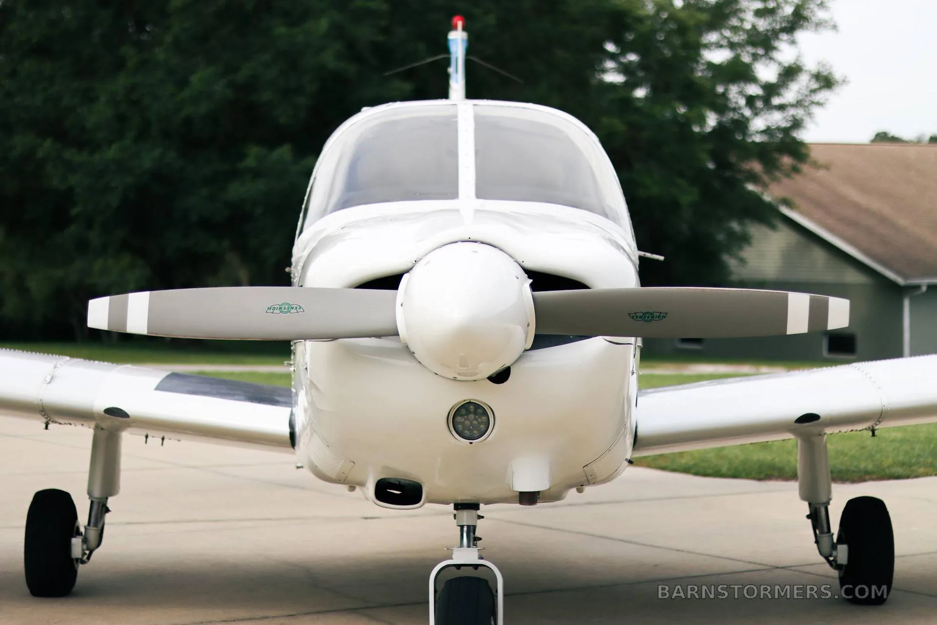 A head-on view of a white Piper PA-28 series aircraft, showing the propeller, nose, and landing gear, parked on a concrete pad.
