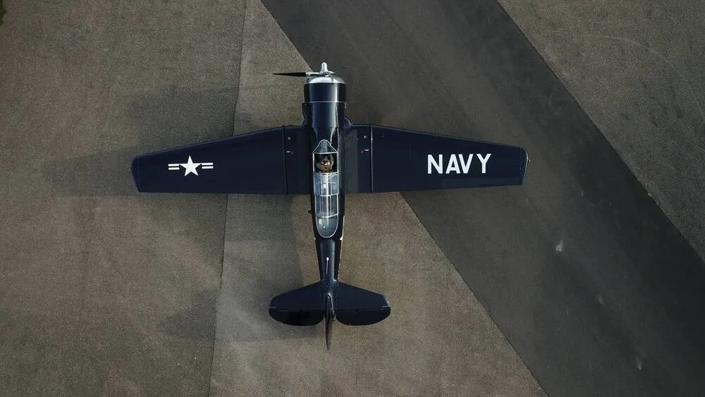 An overhead view of a dark blue North American T-6 Texan aircraft with "NAVY" and a star insignia on its wings, parked on an asphalt surface.