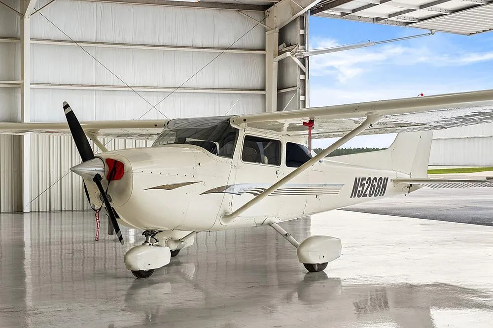 A white Cessna 172, registration N5268H, with gold and gray stripes, parked in a hangar with the door open.