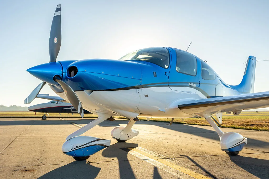A blue and white Cirrus SR22 aircraft parked on an airfield with another aircraft in the background.