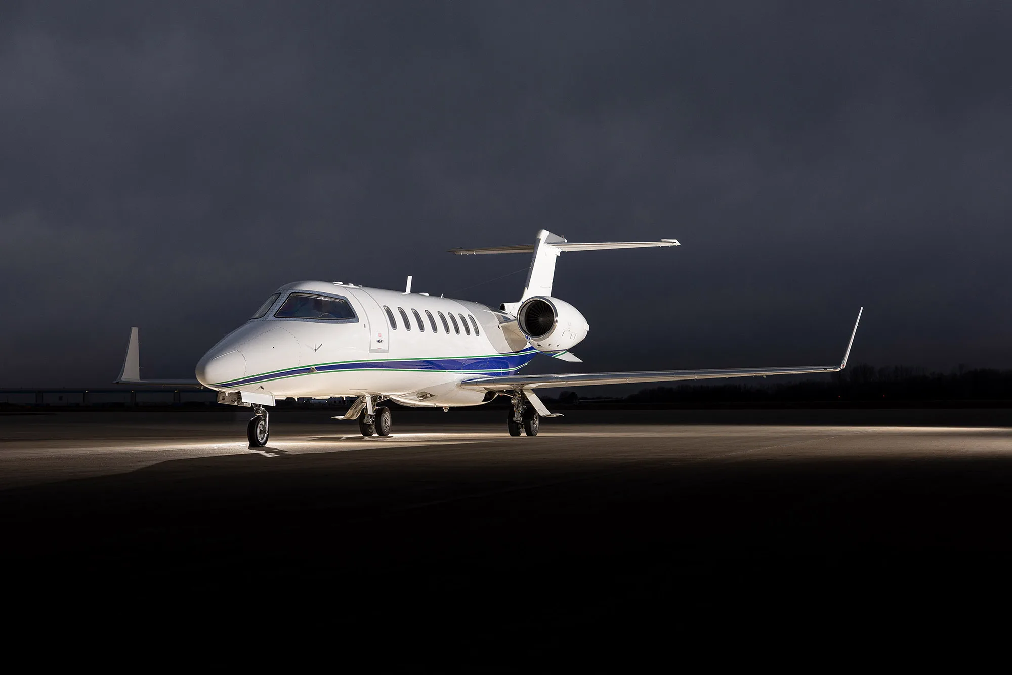 A white Learjet 75 with blue and green stripes parked on an tarmac under dramatic cloudy skies, viewed from the front.