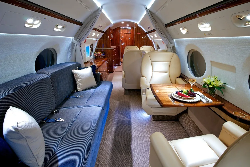 The luxurious cabin interior of a private jet, showing a blue striped sofa, tan leather seats, and a wooden table with a meal.