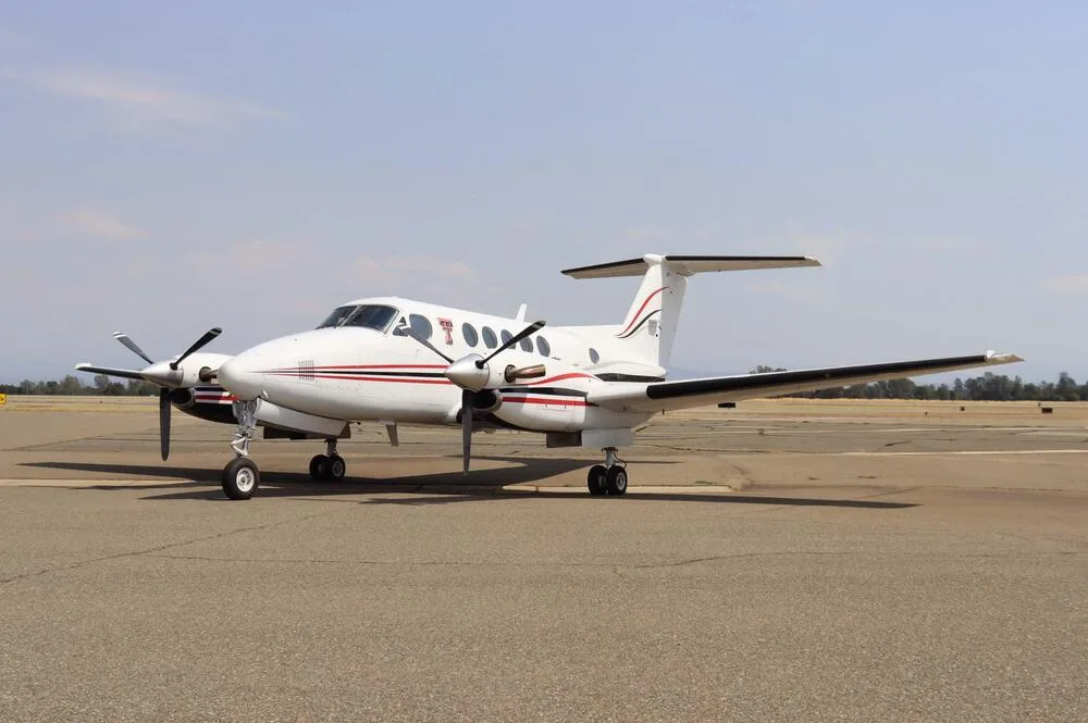 A white Beechcraft King Air with red and black stripes on an airport tarmac, viewed from the front-side.