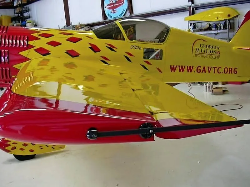 A close-up side view of the yellow and red Sukhoi Su-26 aircraft's fuselage and wing, showing 'GEORGIA AVIATION TECHNICAL COLLEGE' text, inside a hangar.