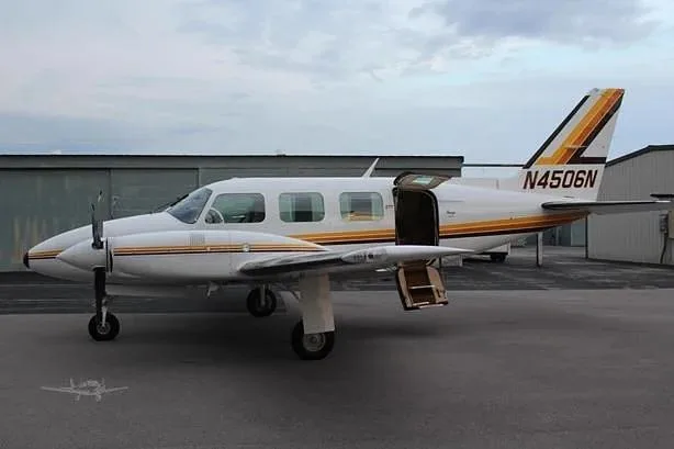 A white twin-engine propeller aircraft with gold and brown stripes, tail number N4506N, is parked on an asphalt surface with its passenger door open.