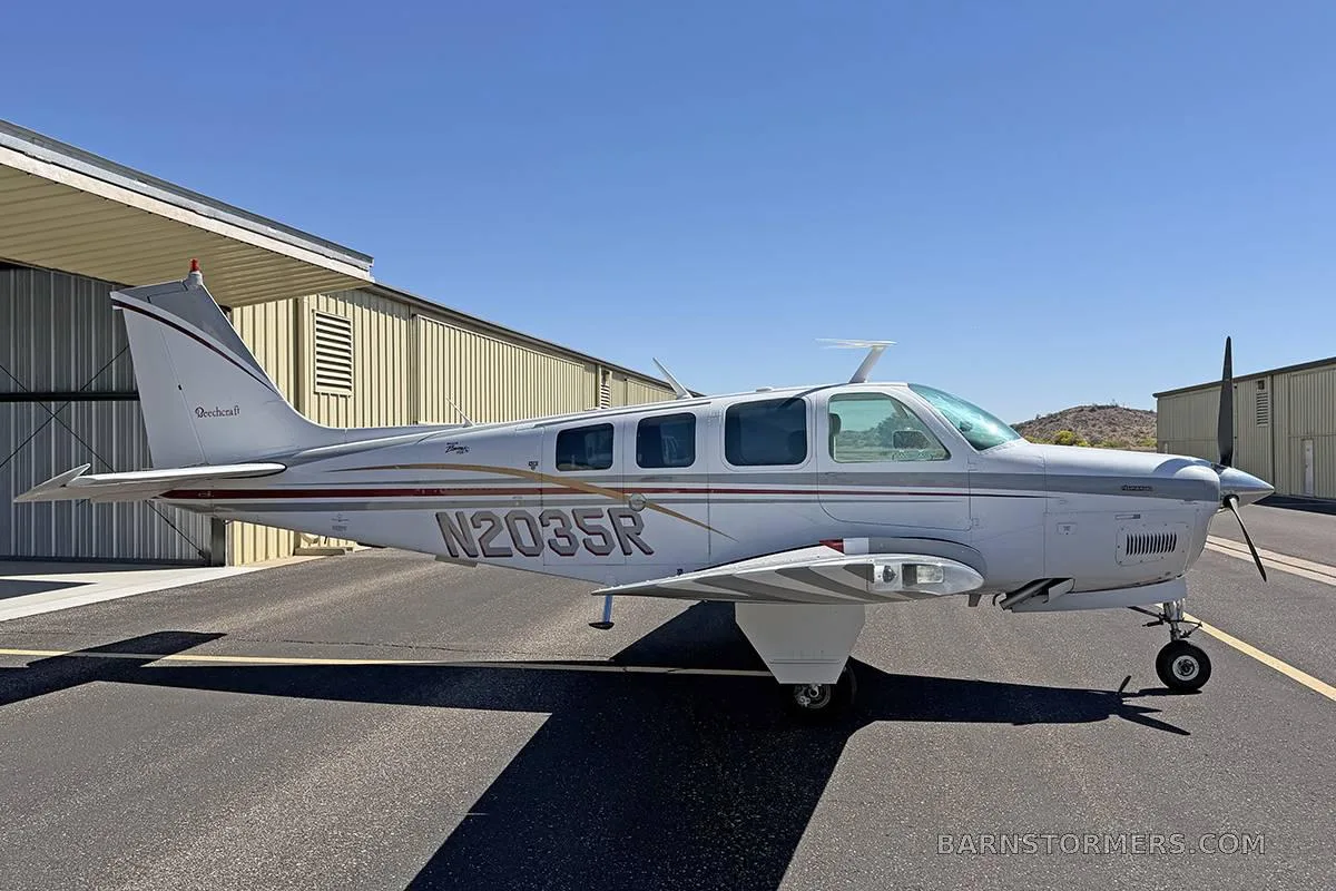 A white Beechcraft Baron with maroon and grey stripes, registration N2035R, parked on an asphalt ramp with a hangar in the background.