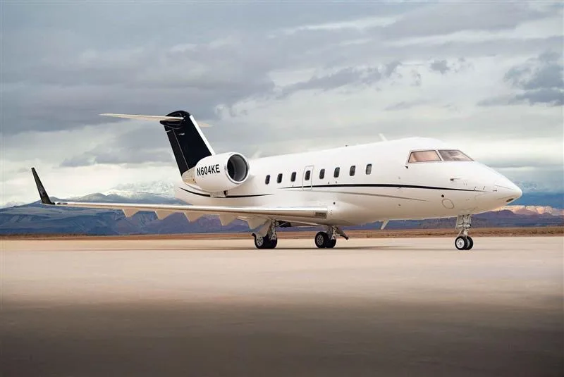 A white Bombardier Challenger 604, registration N604KE, parked on an airfield with a cloudy sky and distant mountains.