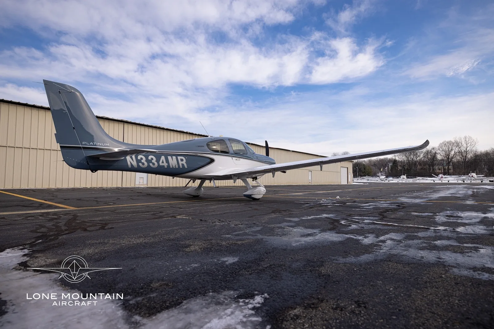 A rear-side view of the Cirrus SR20 aircraft, N334MR, on a snowy tarmac with other aircraft in the distance.