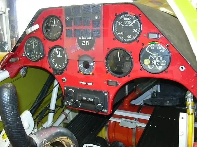 The red instrument panel of a Sukhoi Su-26 cockpit, featuring various analog gauges and a radio.