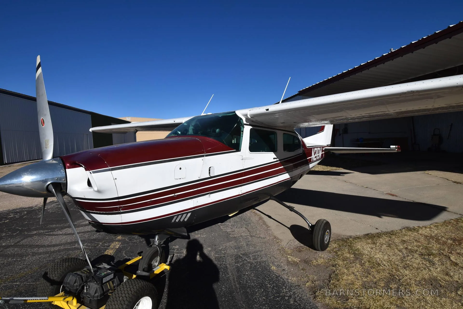 A close-up side view of a white and maroon Cessna 210 aircraft with registration N210W, parked on asphalt next to a hangar.