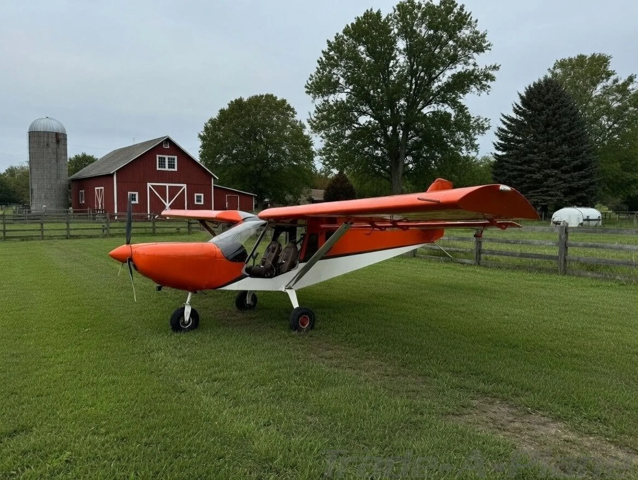 An orange and white light sport aircraft parked on a grassy field in front of a red barn and silo.