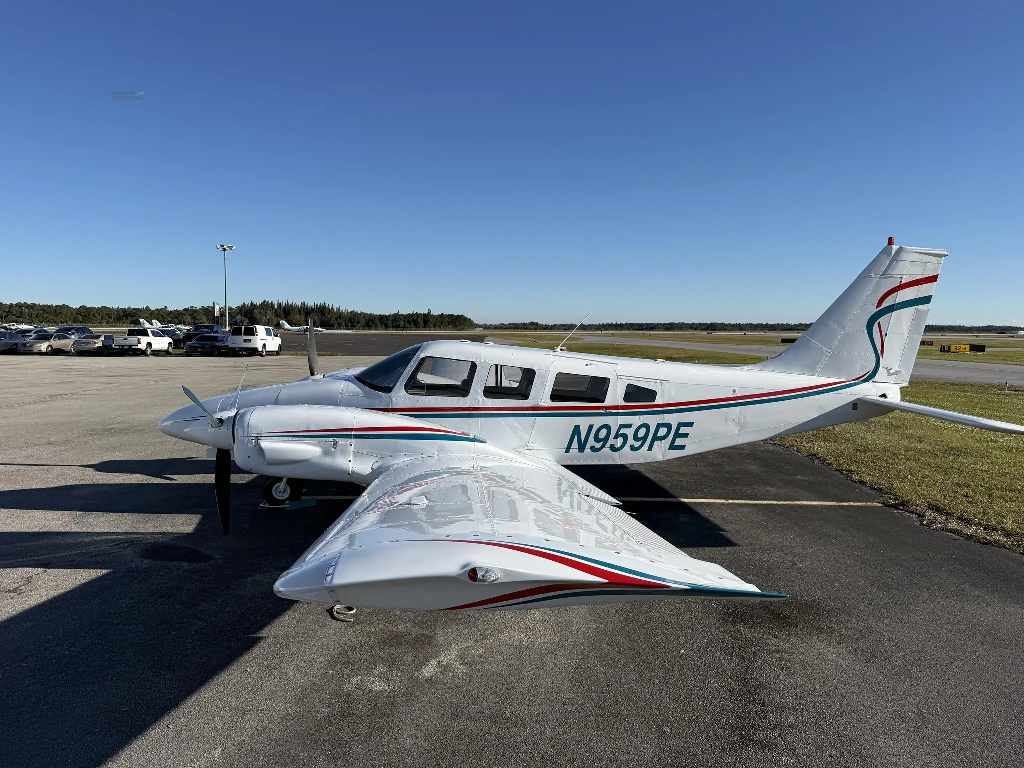 A white twin-engine Piper Seneca II, N959PE, with teal and red stripes, parked on an airport tarmac under a clear sky.