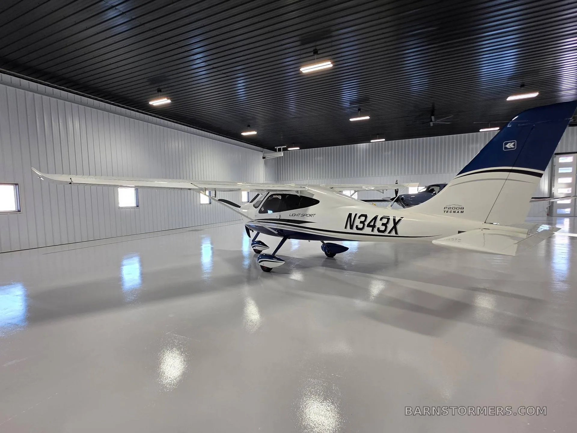 A side view of a white Tecnam P2008 Light Sport aircraft with blue tail and accents, registration N343X, parked in a hangar.