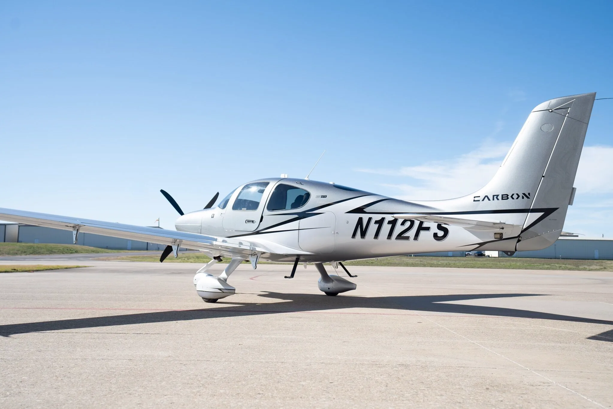 A side-rear view of a silver and dark grey Cirrus SR22T Carbon Edition aircraft (N112FS) parked on an airport tarmac.