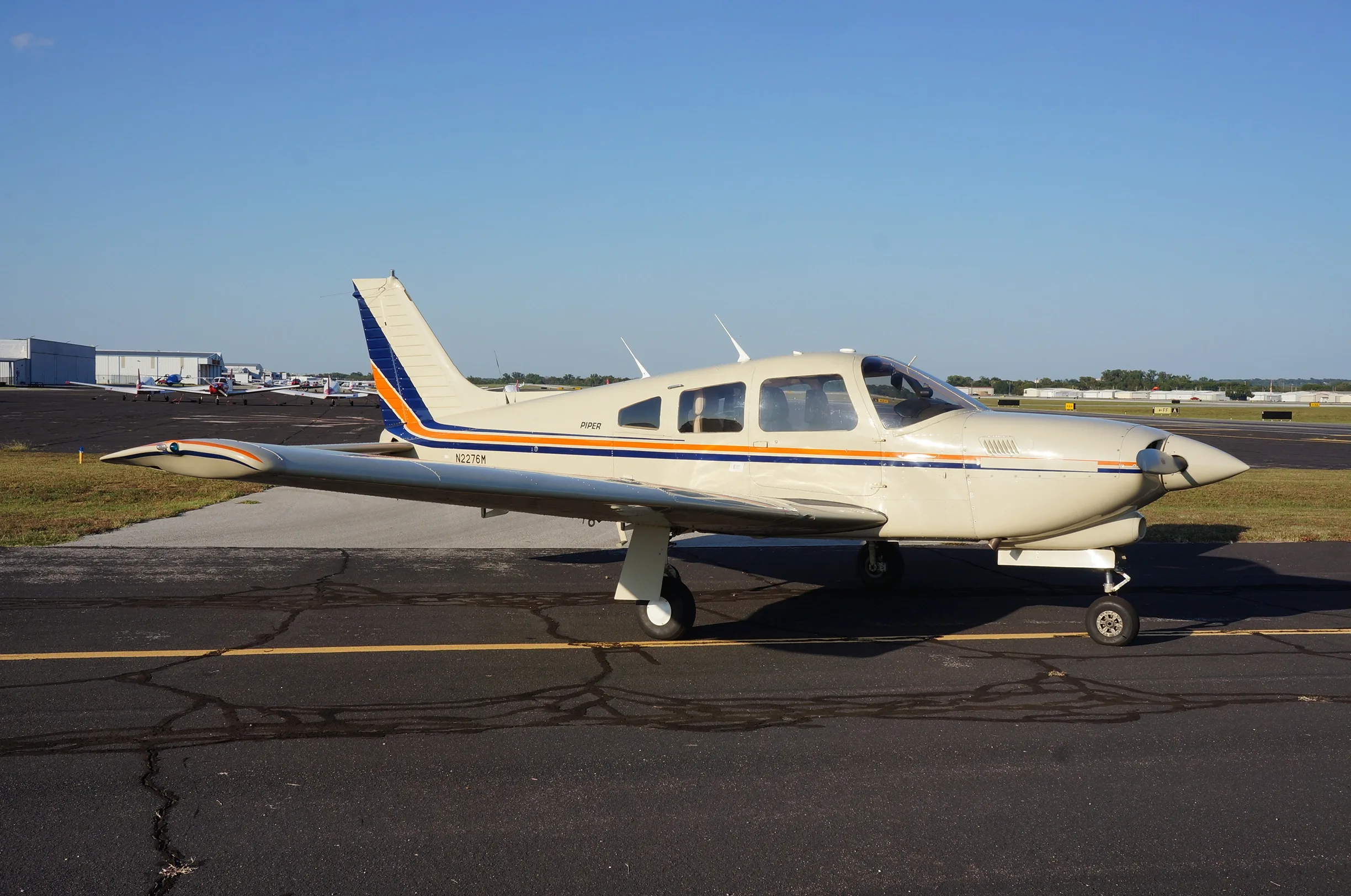 A cream-colored Piper aircraft, registration N2276M, with blue and orange stripes, parked on an asphalt tarmac.