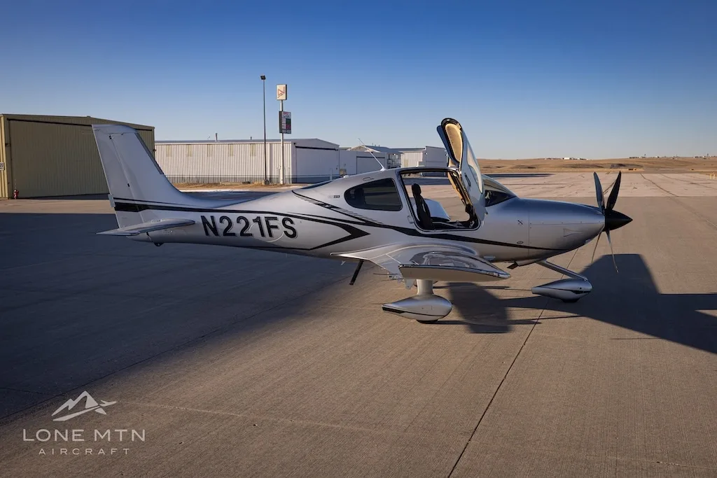 A side view of the silver Cirrus SR22T aircraft N221FS with its doors open, parked on an airfield.