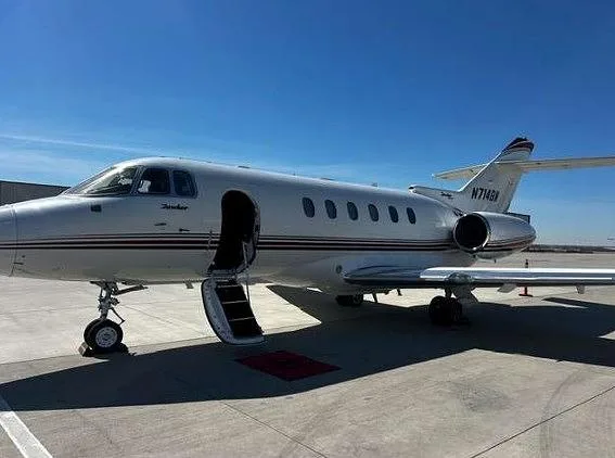 A white Hawker business jet with red and black stripes, tail number N714BW, parked on an airfield with its main cabin door open.