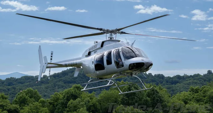 A white Bell 206 JetRanger helicopter with registration N110JJ flying over green treetops under a blue sky.