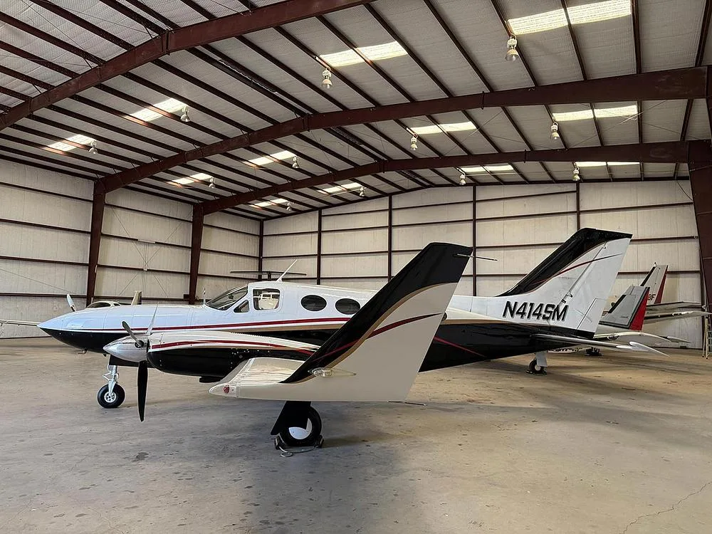 A white and black twin-engine aircraft, N414SM, with gold trim, parked inside a hangar, viewed from the rear-left.