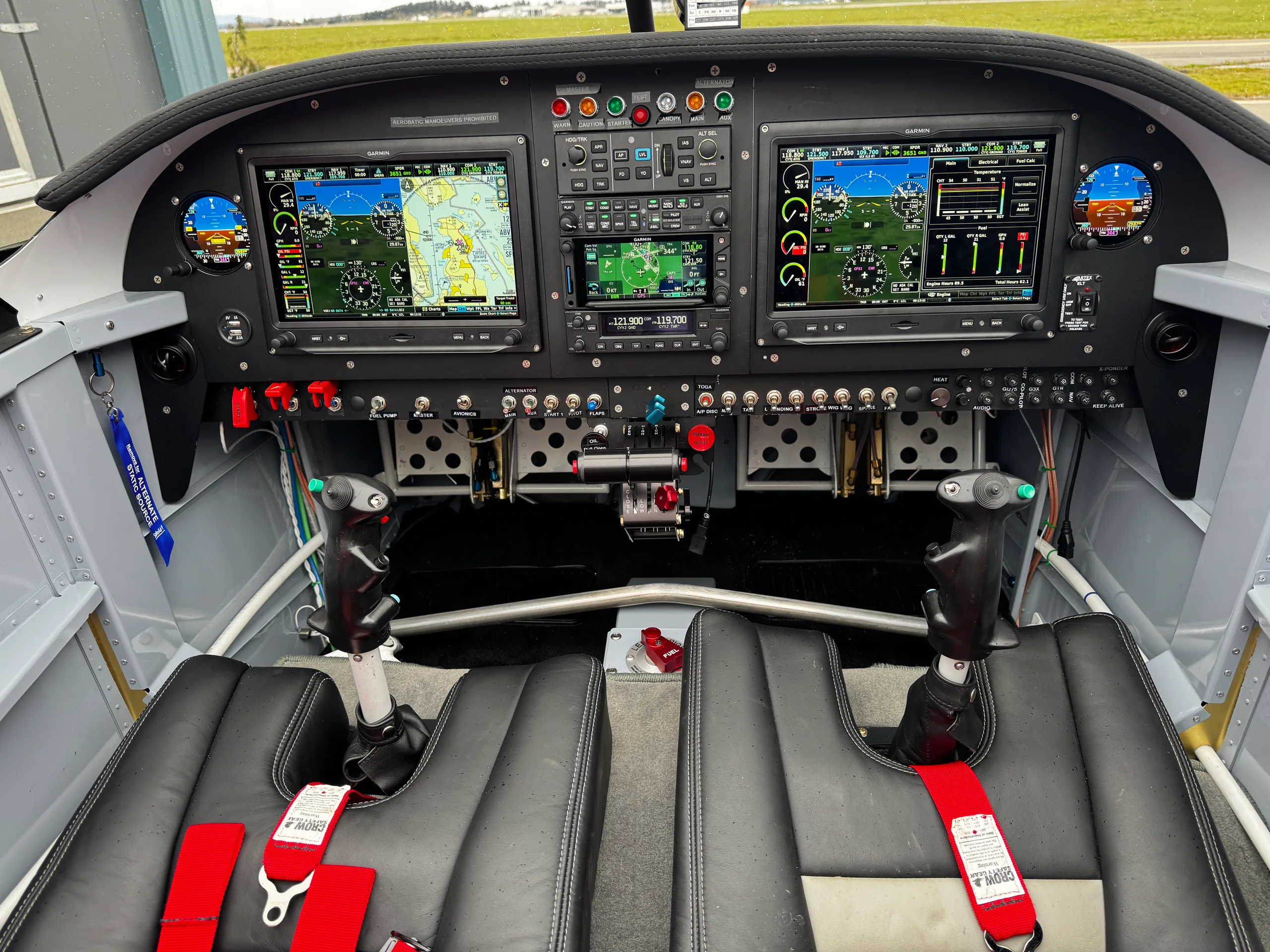 The cockpit of a small aircraft, featuring two large Garmin displays, a central control panel, and two black seats with red harnesses.