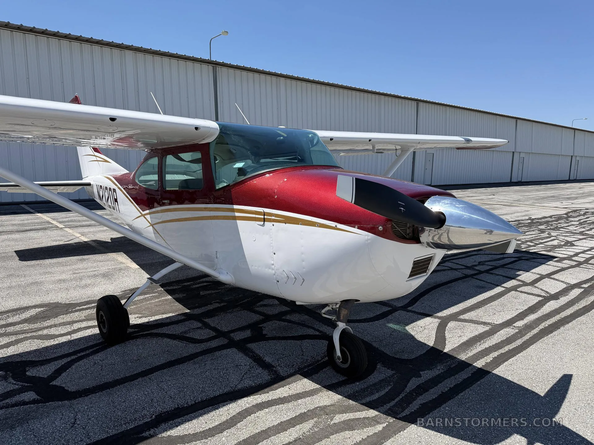 A front-quarter view of a red and white Cessna 182 aircraft with a polished propeller spinner parked on an asphalt apron.
