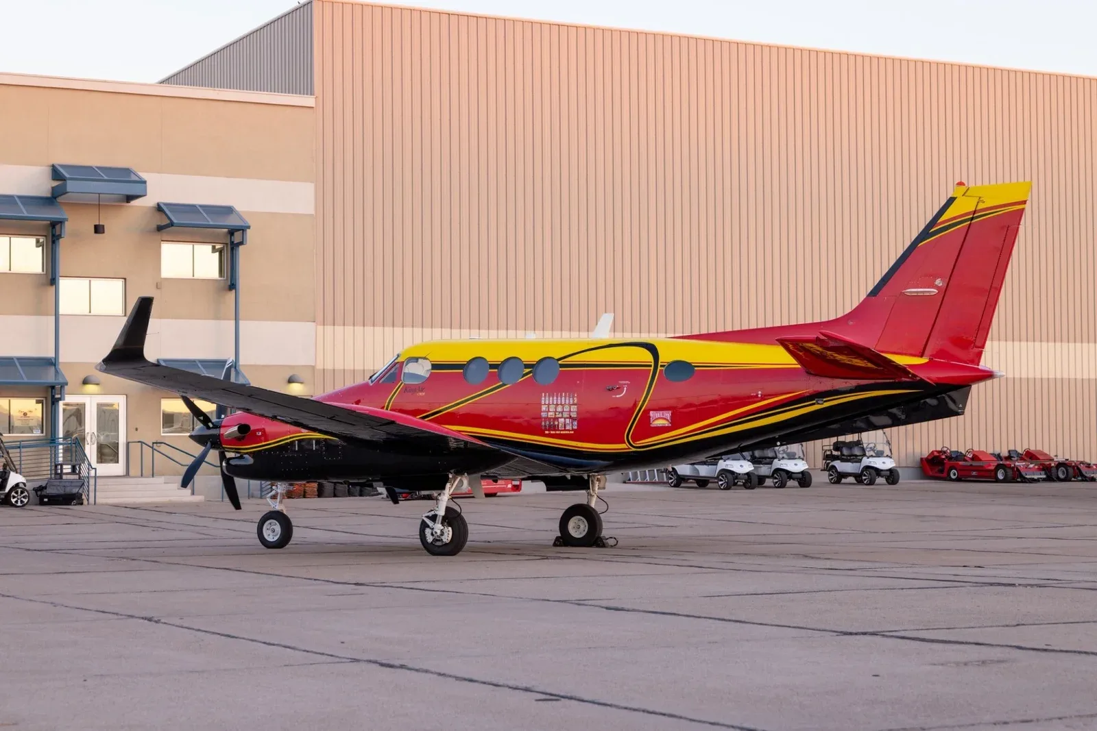 A red, yellow, and black King Air C90B turboprop aircraft parked on an apron next to a building.