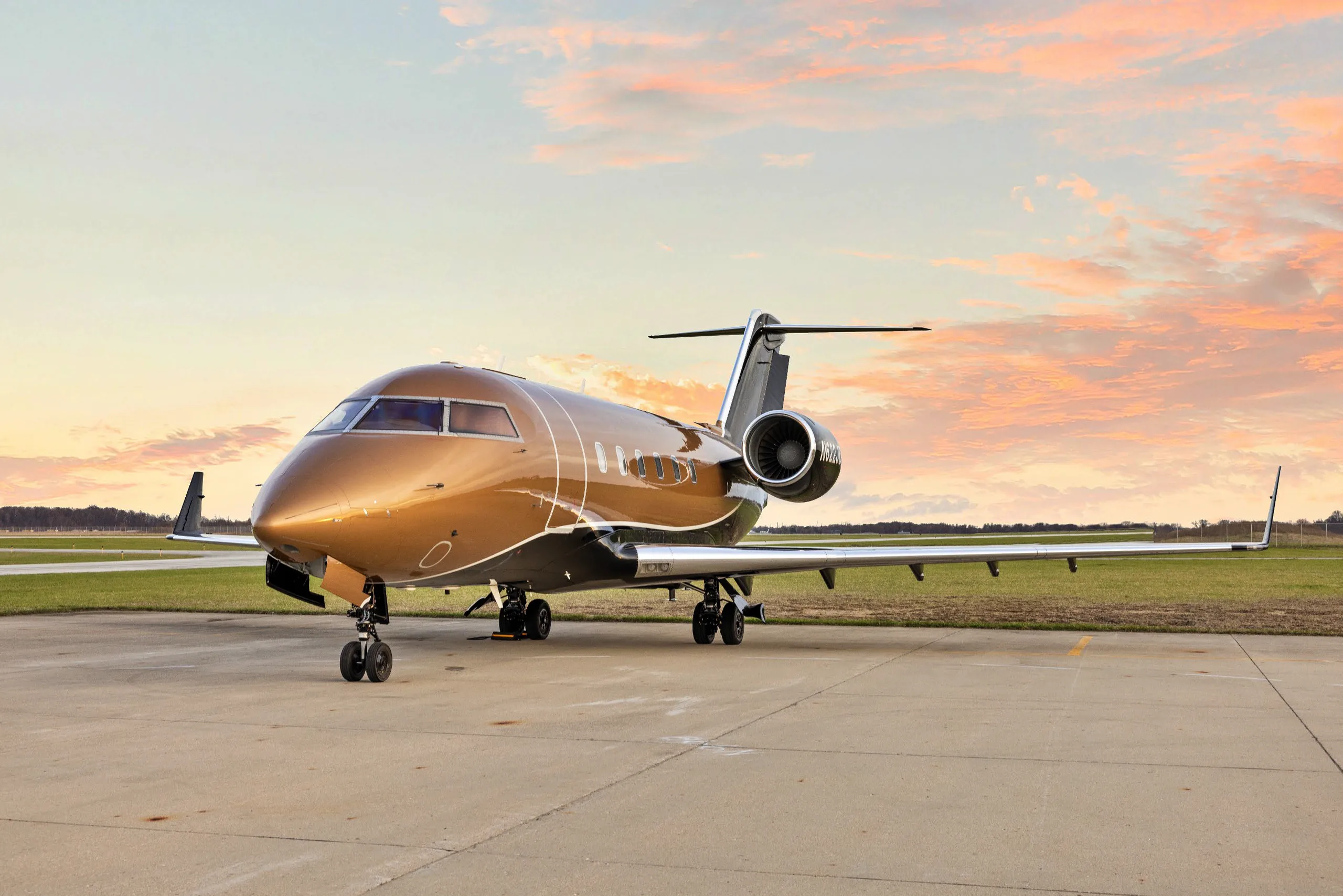 A gold and black Bombardier Challenger private jet is parked on an airport tarmac under a sunset sky.