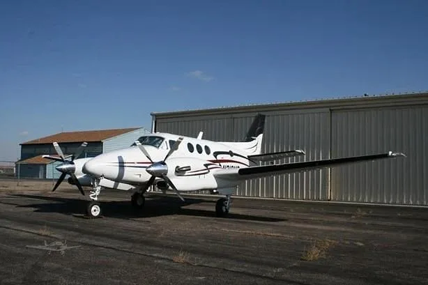 A white twin-engine turboprop aircraft with black and grey stripes, parked on an asphalt ramp in front of a metal hangar under a clear blue sky.