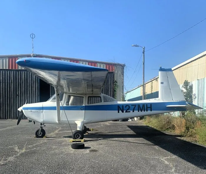 A white and blue high-wing aircraft, registration N27MH, is parked on an asphalt apron with hangars in the background.
