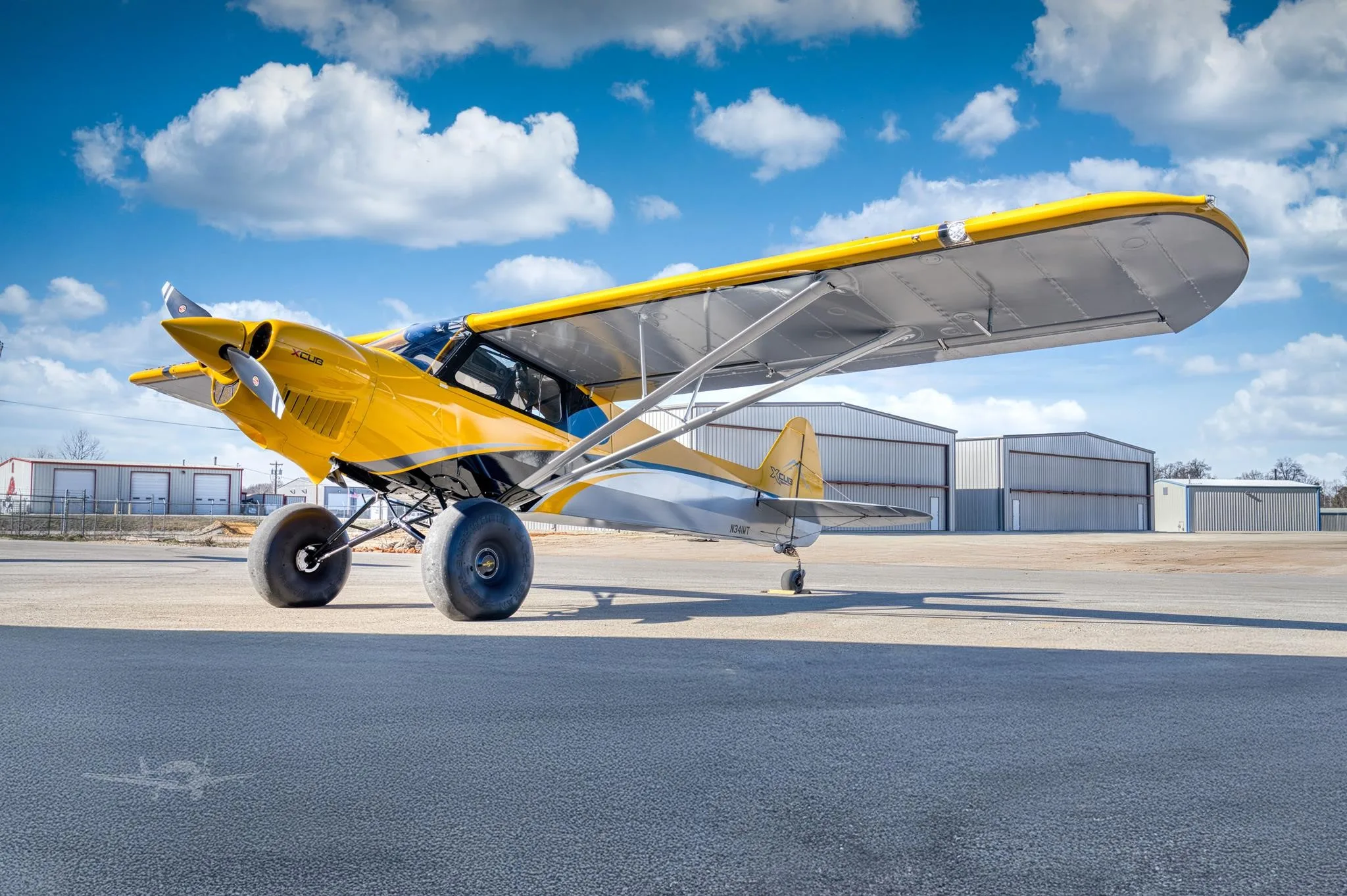 A yellow and grey X-Cub aircraft with large tundra tires is parked on an airfield apron with hangars in the background under a blue sky.