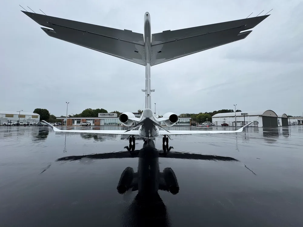 Rear view of a white business jet with two engines, parked on a wet tarmac with its reflection visible.