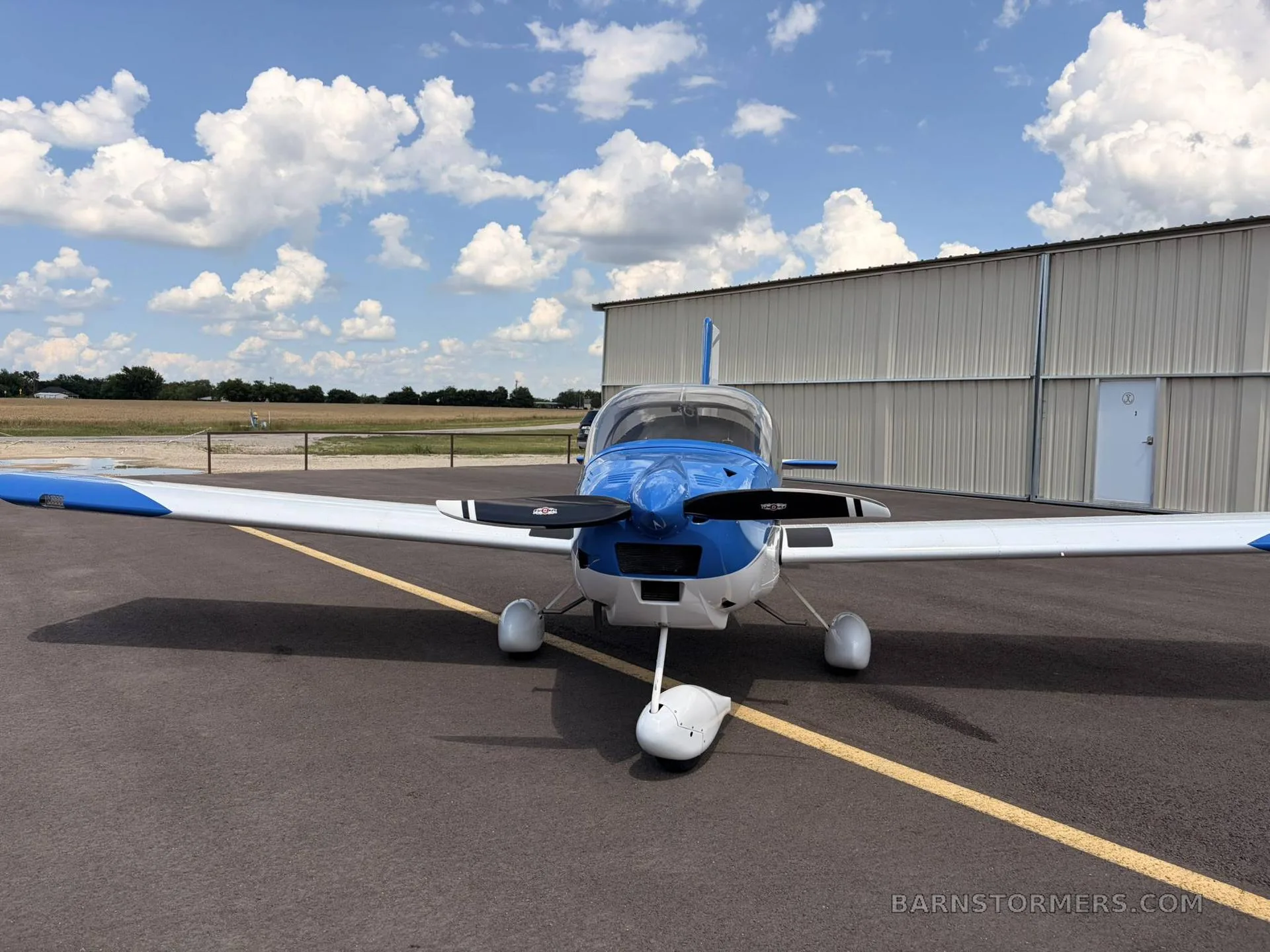 A blue and white single-engine aircraft with a propeller, parked on an asphalt tarmac in front of a hangar under a partly cloudy sky.