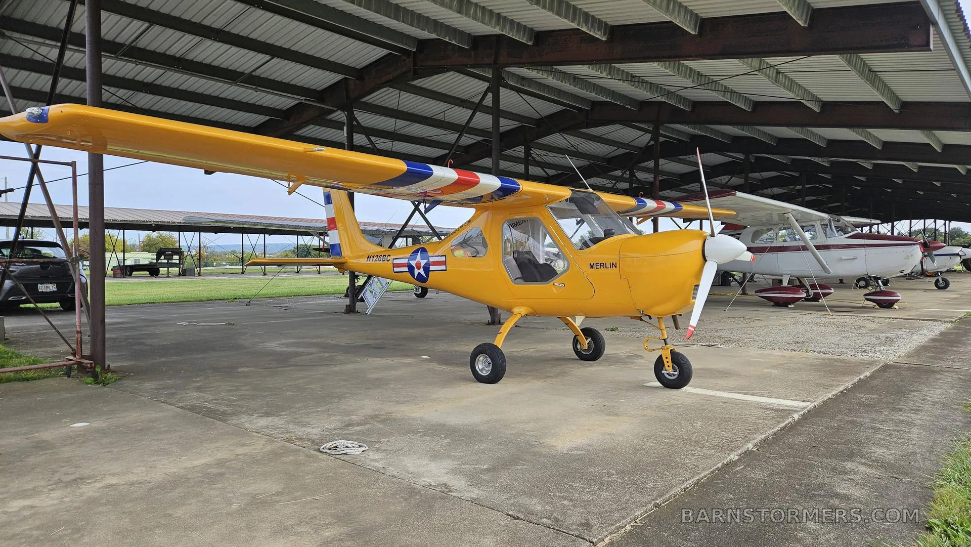 A yellow Merlin ultralight aircraft with a red, white, and blue striped wing is parked under an open-sided hangar.