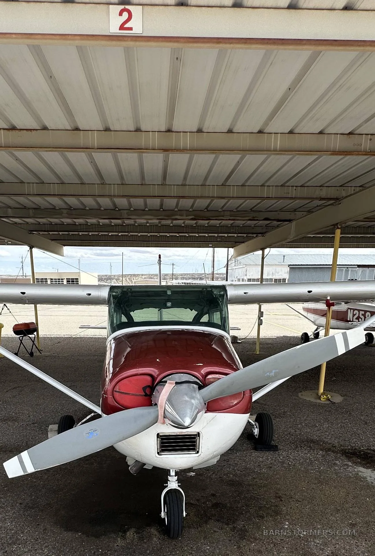 A front view of a red and white single-engine propeller aircraft (Cessna) parked under a metal shelter.