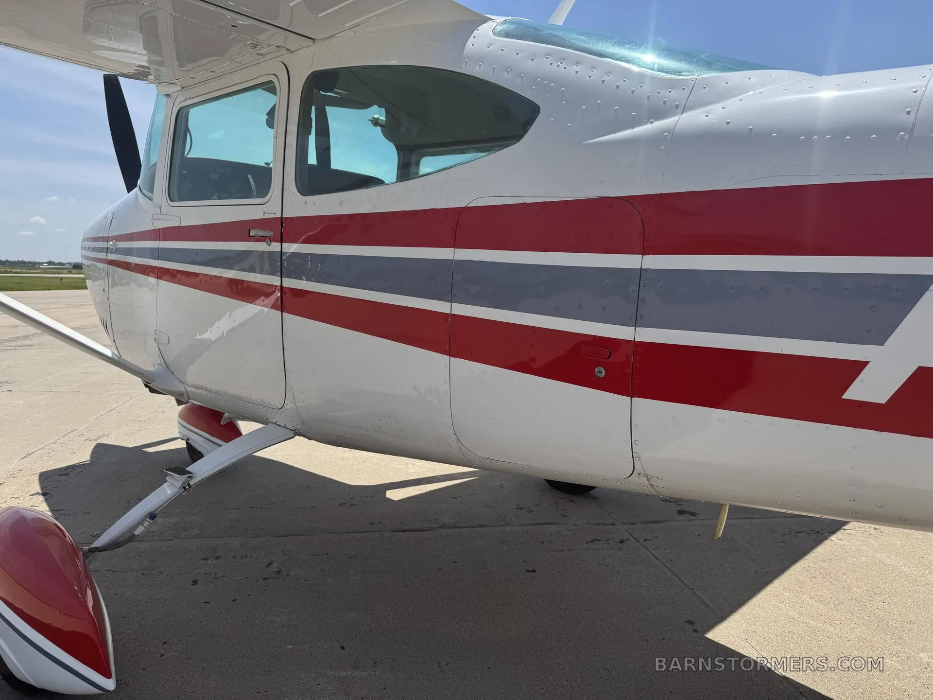 A close-up side view of the white fuselage of a Cessna aircraft with red and grey stripes, showing the cabin windows and propeller.