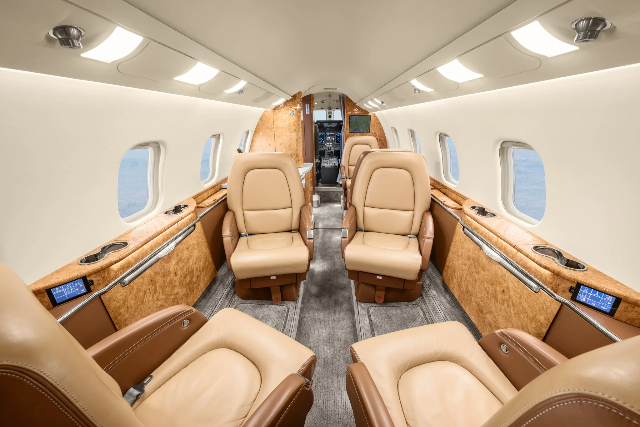 The spacious interior cabin of a private jet, showing rows of tan leather seats and wood paneling, looking towards the cockpit.