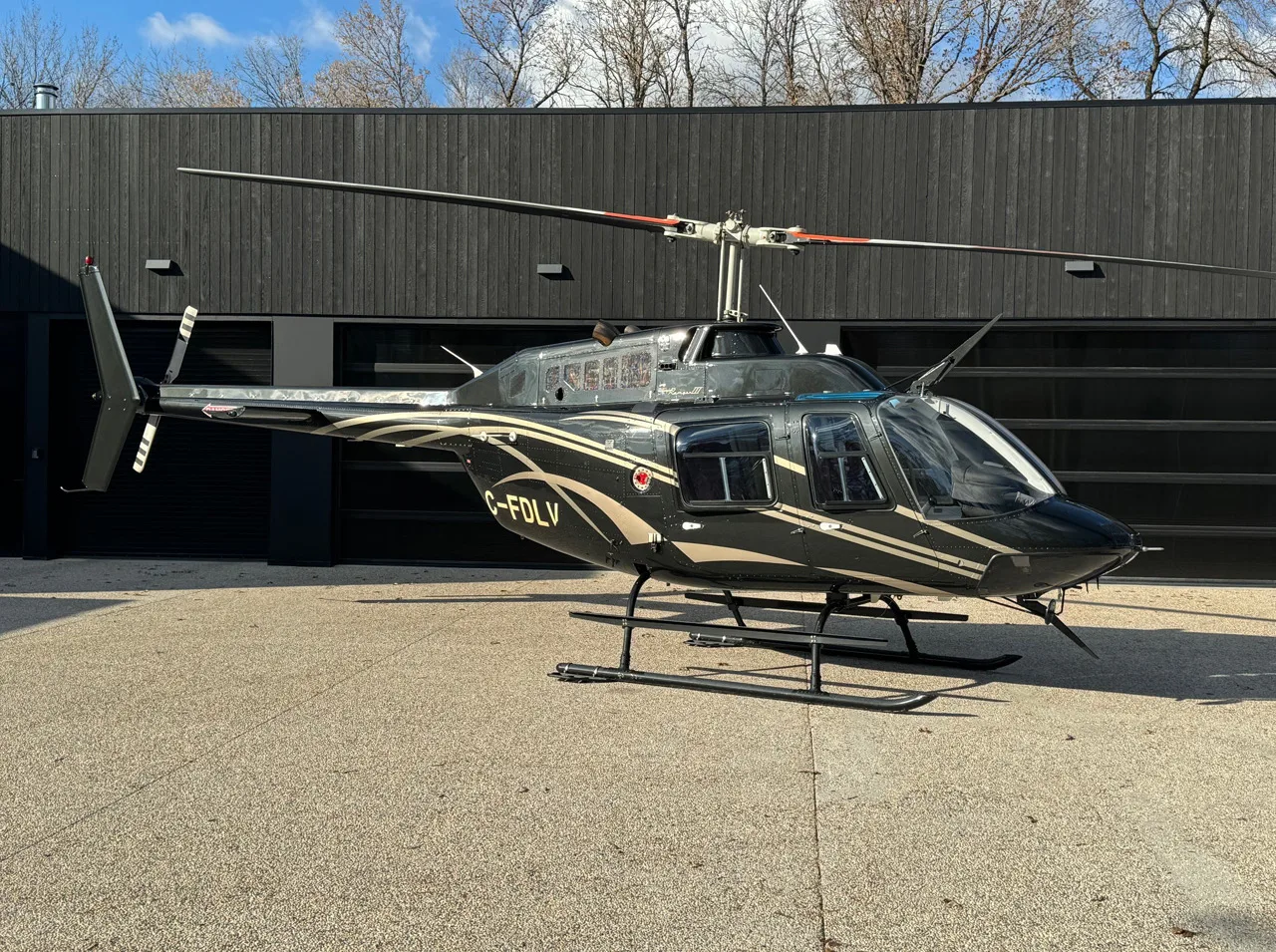 A dark green Bell 206B JetRanger III helicopter with gold stripes is parked on a concrete pad in front of a modern black building.