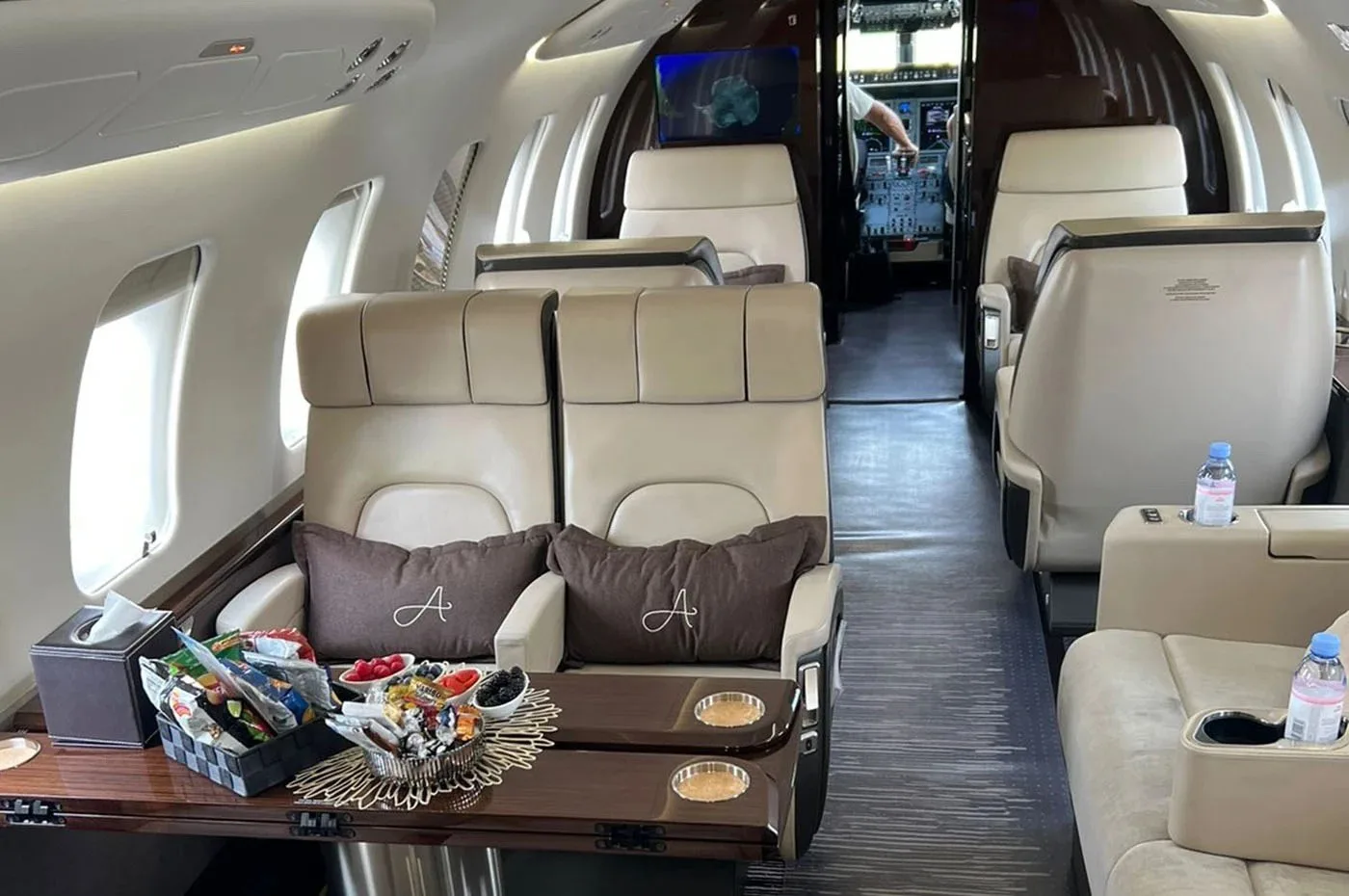 The private jet's cabin with beige seats, a table with snacks, and a view into the cockpit.