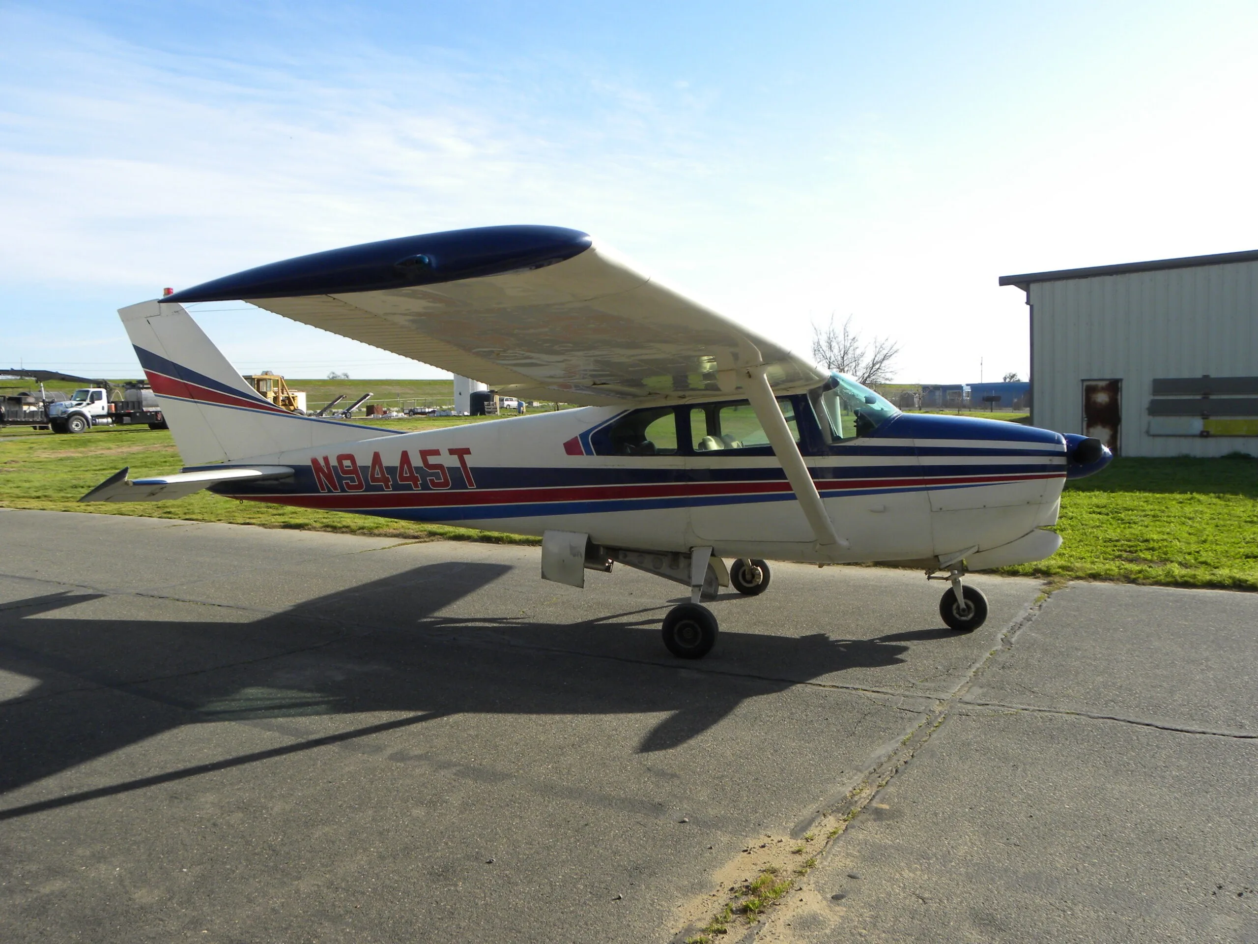 A side view of a white Cessna 182 with blue and red stripes, registration N9445T, parked on a tarmac.
