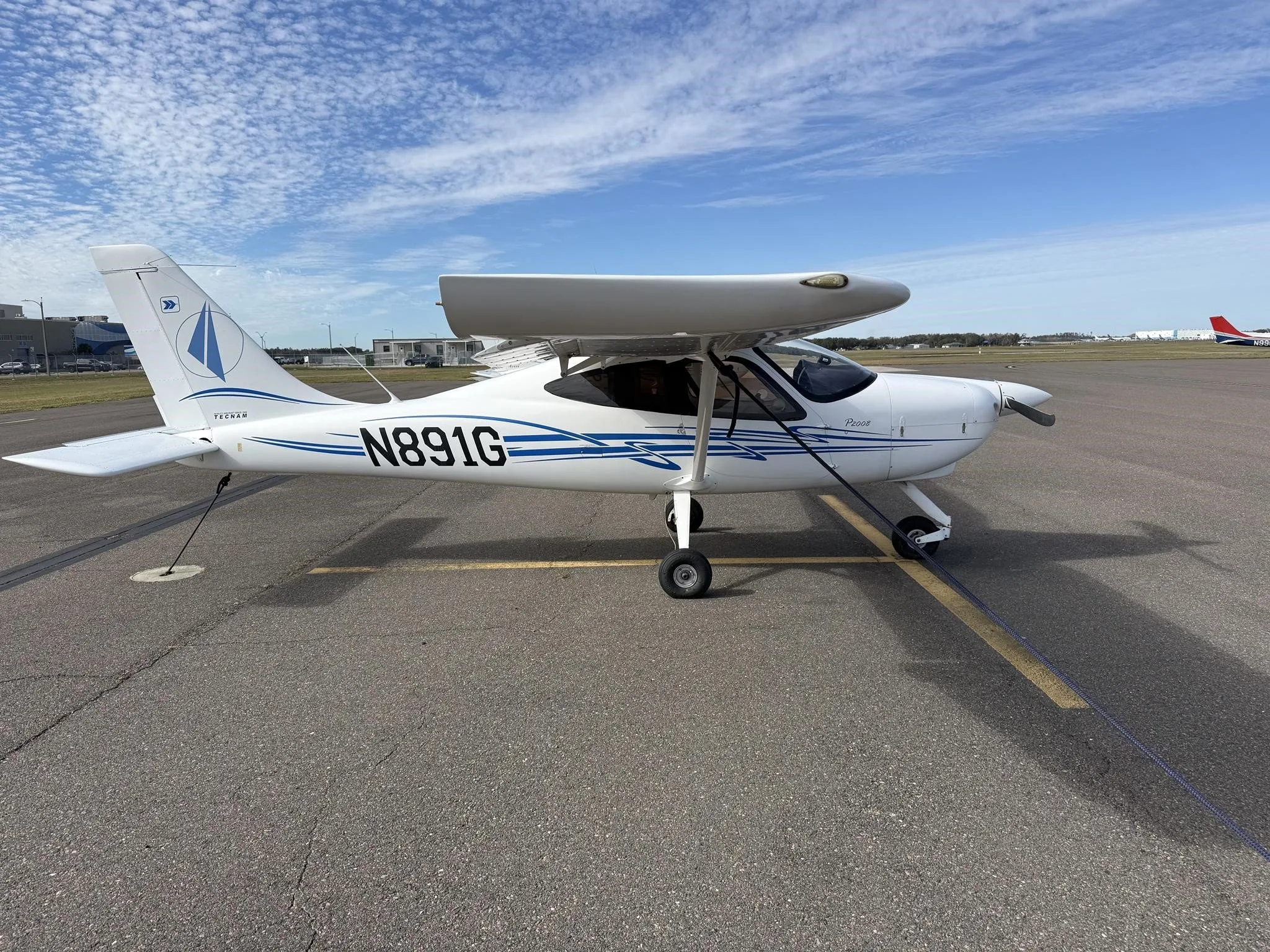 A white Tecnam P2008 aircraft with blue stripes, registration N891G, parked on an airport tarmac with wing covers on.