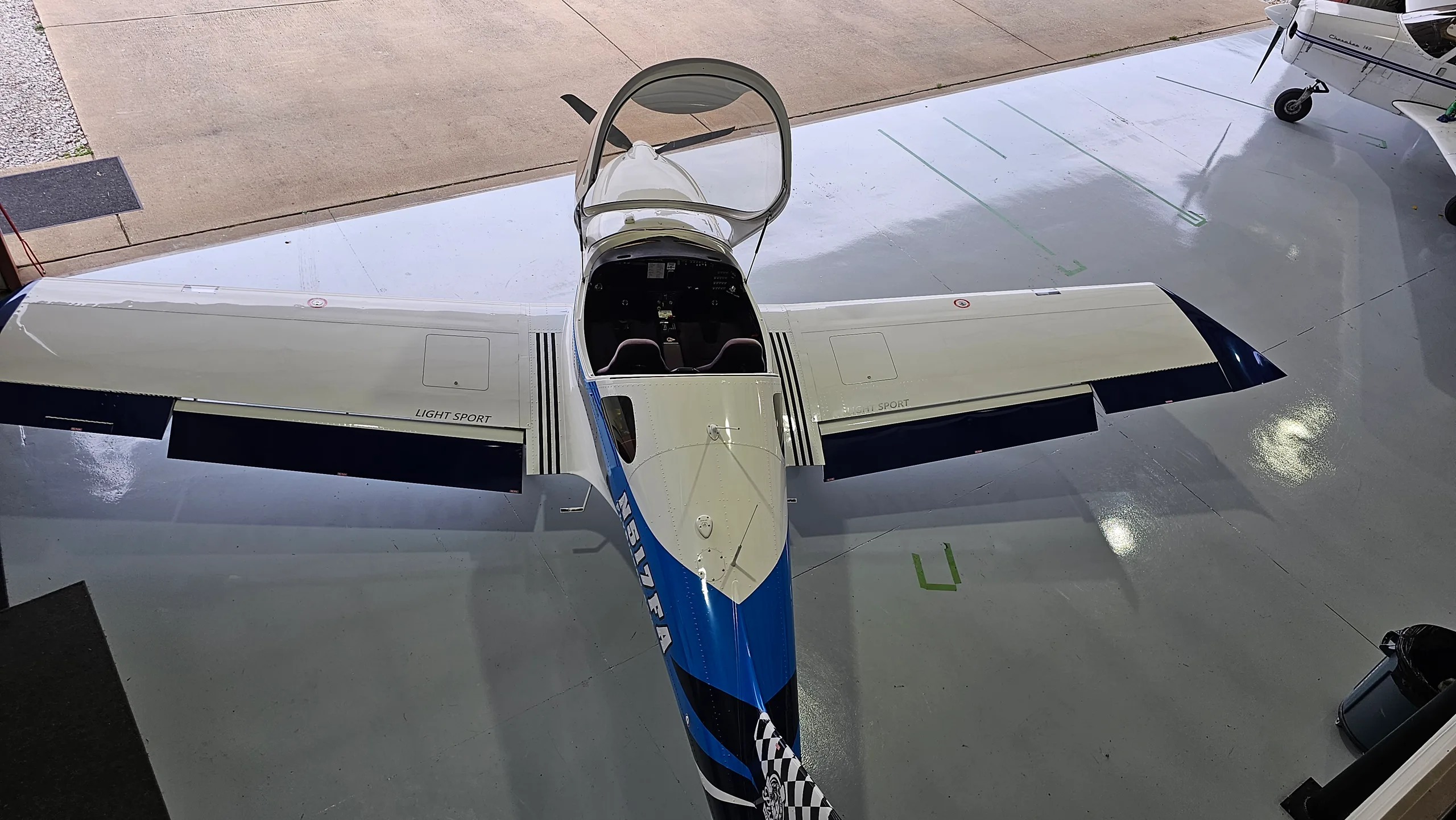 A top-down view of a white and blue light sport aircraft with an open canopy, parked in a hangar.