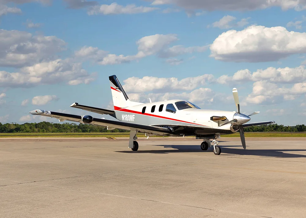 A white and grey TBM 900 series aircraft with red stripes, registration N180MF, parked on an airfield under a partly cloudy sky.
