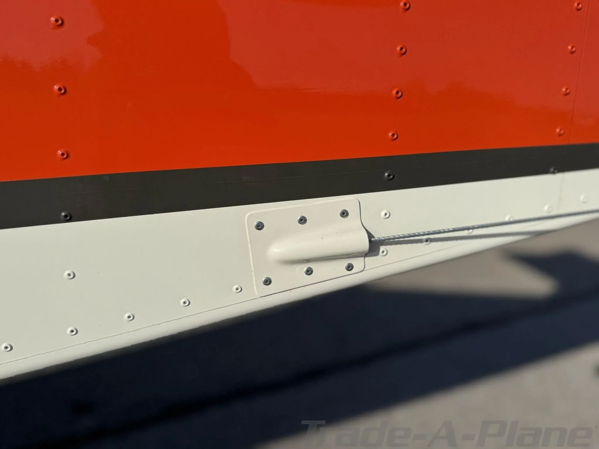 Close-up view of the orange and white fuselage of an aircraft, showing rivets and a control cable fairing.