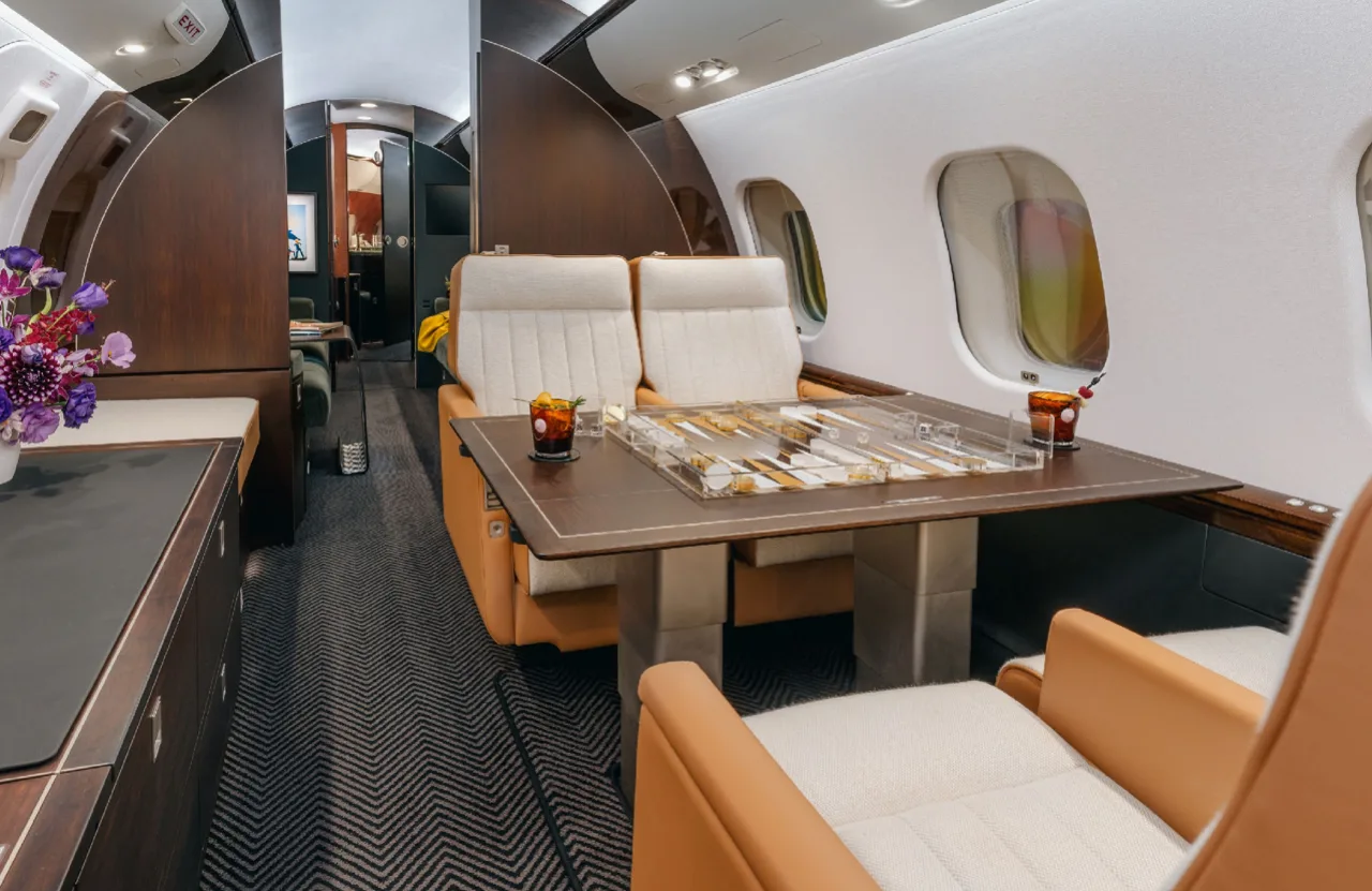 A wide view of the private jet's luxurious cabin, showing cream and tan seats, a table with a backgammon set, and a galley area in the background.