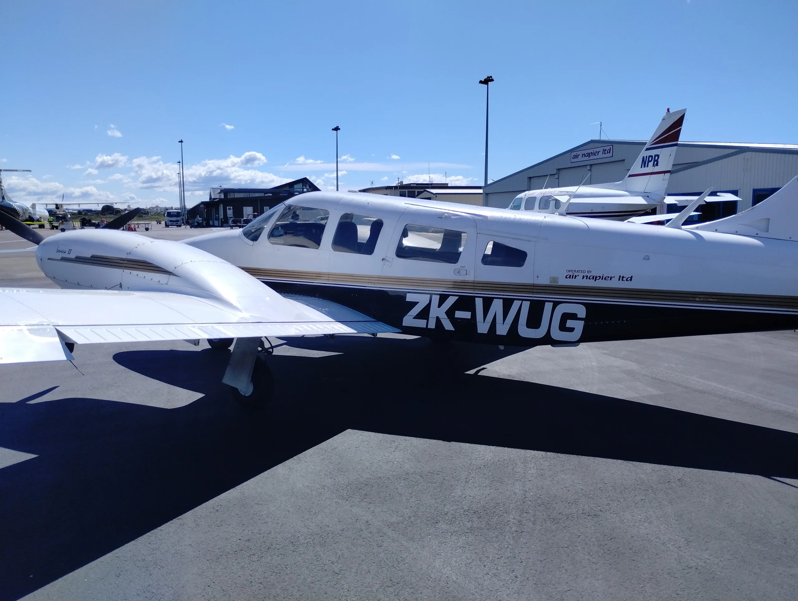 A white and black Piper Seneca II aircraft with registration ZK-WUG parked on an airfield next to hangars.