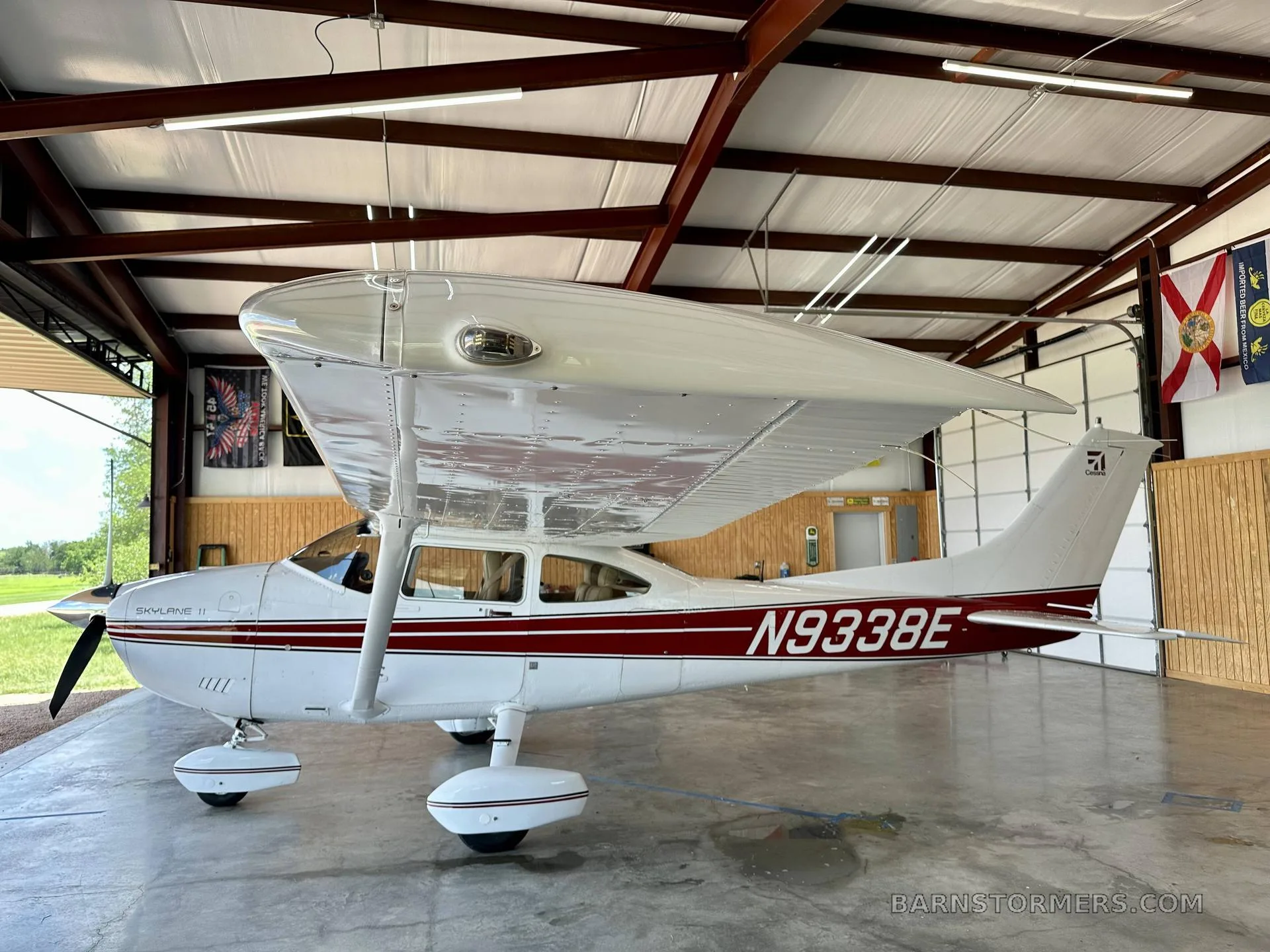 A white Cessna Skylane II with red stripes and registration N9338E is parked inside a hangar, viewed from the rear-left.