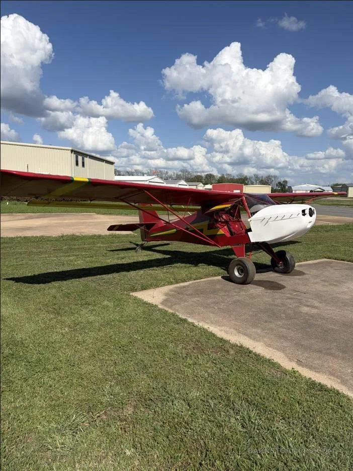 A red and yellow ultralight aircraft with a white nose cowl parked on grass next to a concrete pad under a blue sky with clouds.